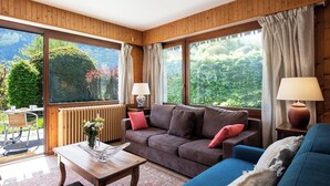 Una stanza con soffitto in legno boiserie, un divano marrone, un divano blu, un tavolo in legno e vista sul verde attraverso ampie finestre.