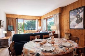 A dining area with a table set for two, a sofa, and a television in a room with wooden walls and large windows.