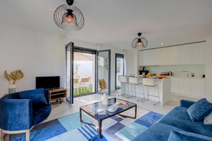 A modern living room with a glass coffee table, blue sofas, a TV, and a kitchen area with a bar.