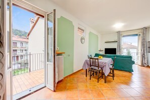 A room with a dining table, chairs, a green sofa, a TV, and a balcony with a view of buildings and a hill.