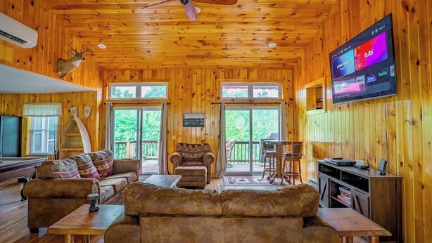 A cozy cabin interior with a wooden ceiling, a large TV mounted on the wall, a sofa set, and a fireplace. - Spacious 5 Bedroom Cabin Riverfront/Black Lake Access (Hammond)