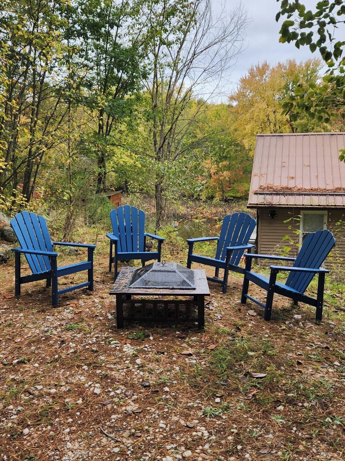 Uma área de estar ao ar livre rústica, com quatro cadeiras Adirondack e uma mesa quadrada, cercada por árvores e um pequeno edifício com telhado de metal.