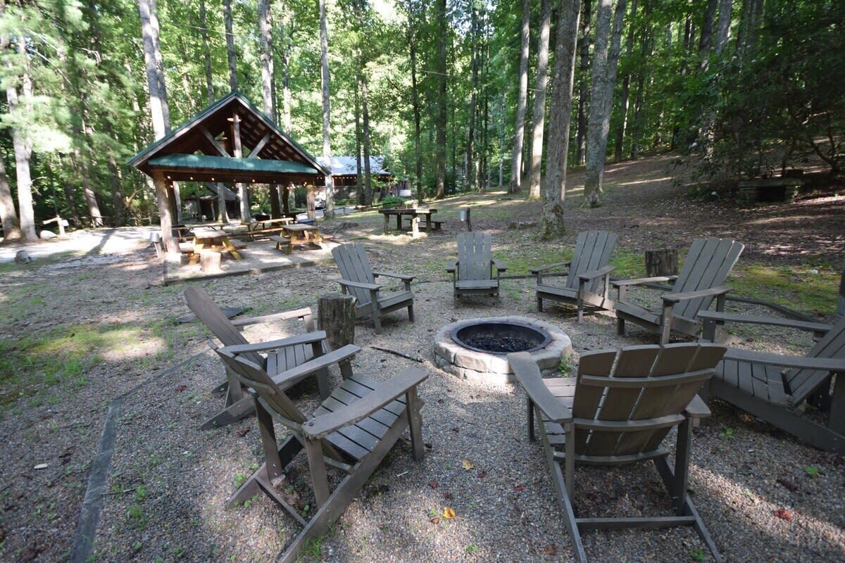 Un camping con sillas Adirondack de madera dispuestas alrededor de un brasero central, un refugio cubierto para picnic y mesas de picnic.