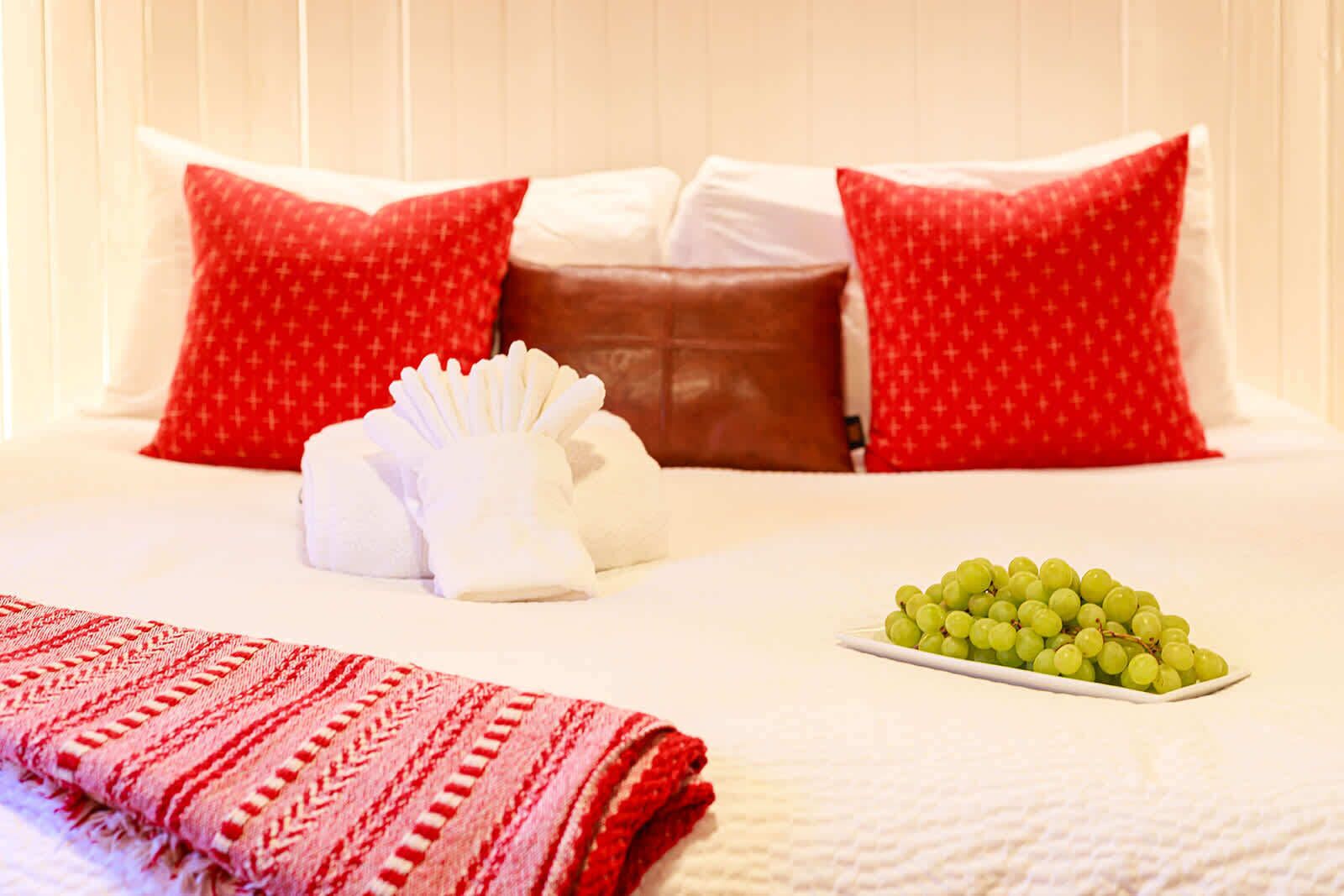 A hotel room with a neatly made bed, red patterned pillows, a white towel, and a plate of green grapes.