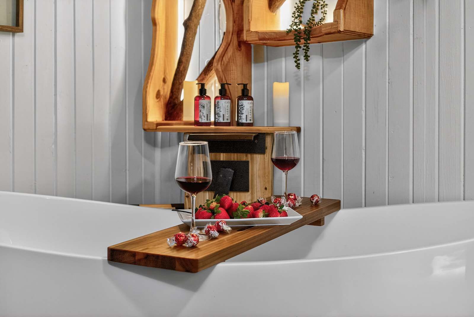 Una bandeja de madera con fresas y copas de vino sobre una superficie blanca, con botellas y velas al fondo.
