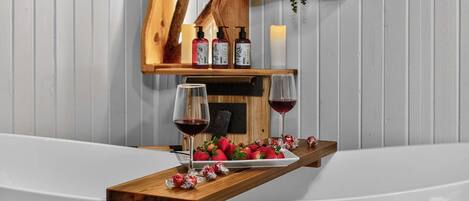 Una bandeja de madera con fresas y copas de vino sobre una superficie blanca, con botellas y velas al fondo.