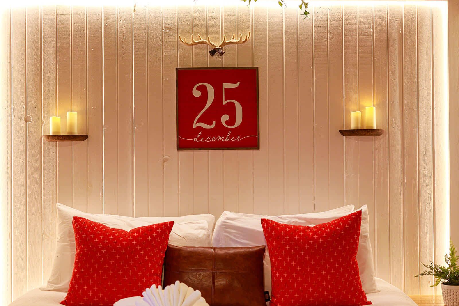 A bed with white and red pillows, a red sign with '25 December', and wall-mounted candles.