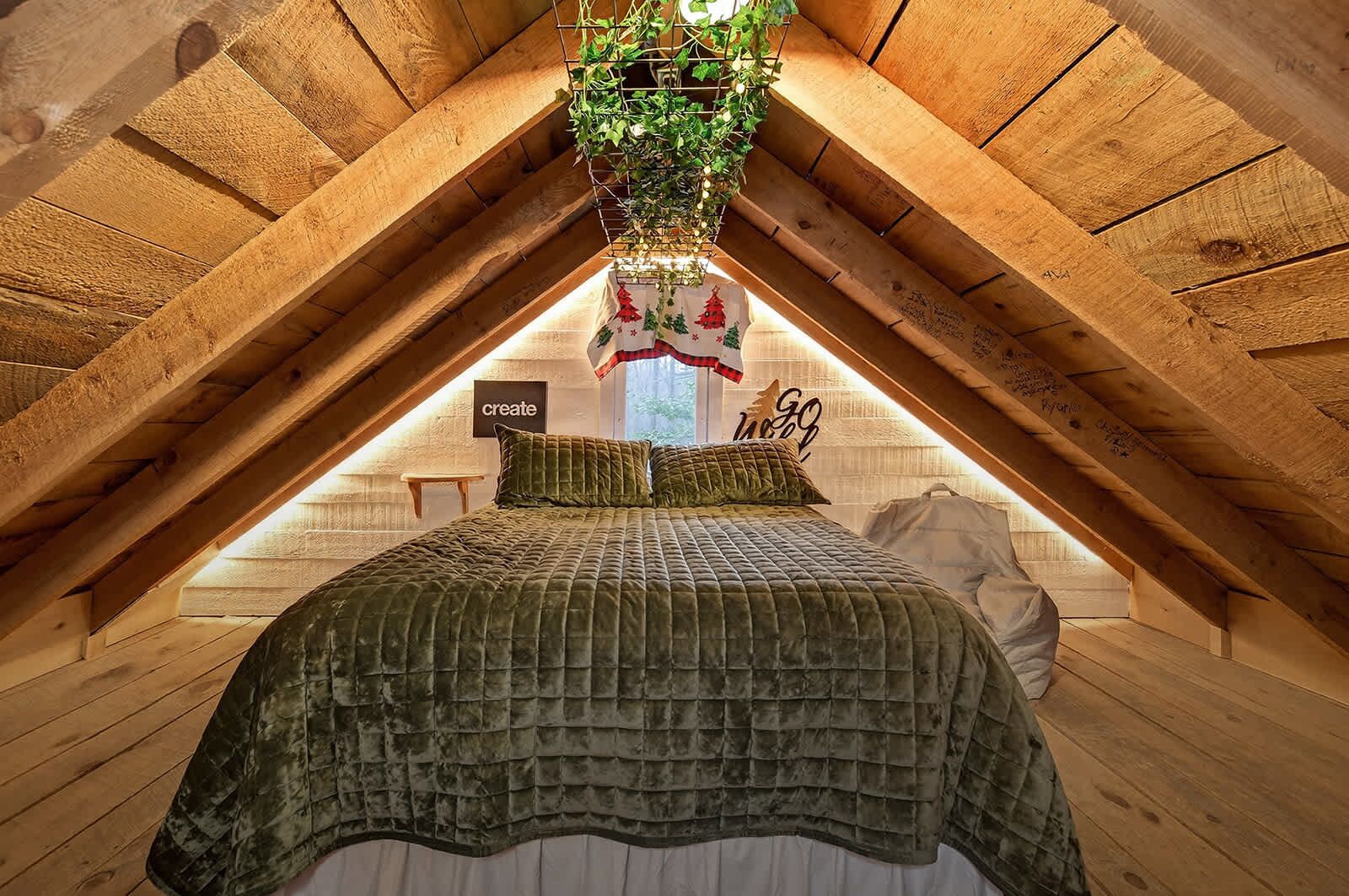 Une chambre mansardée chaleureuse, avec un plafond en pente, un lit recouvert d’une couette verte, une petite table et une plante suspendue.