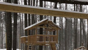 Un parque infantil de madera con un suelo cubierto de nieve y una casita de juegos de madera en un bosque nevado.