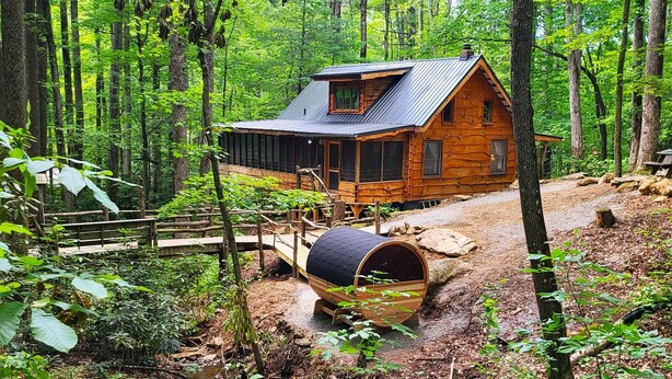 Une cabane rustique, dotée d’un toit en pente, est entourée d’une forêt dense. Un tonneau en bois est placé devant la cabane, et un pont en bois y mène.