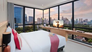 Ein modernes Hotelzimmer mit einem großen Bett, einem Fernseher auf einer Holzhilfe und Blick auf die Stadt durch große Fenster.