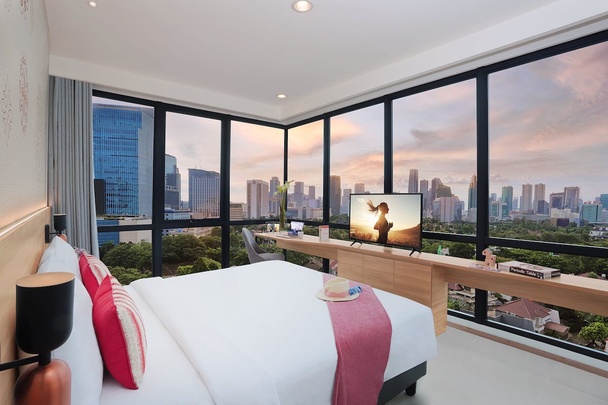 Ein modernes Hotelzimmer mit einem großen Bett, einem Fernseher auf einer Holzhilfe und Blick auf die Stadt durch große Fenster.