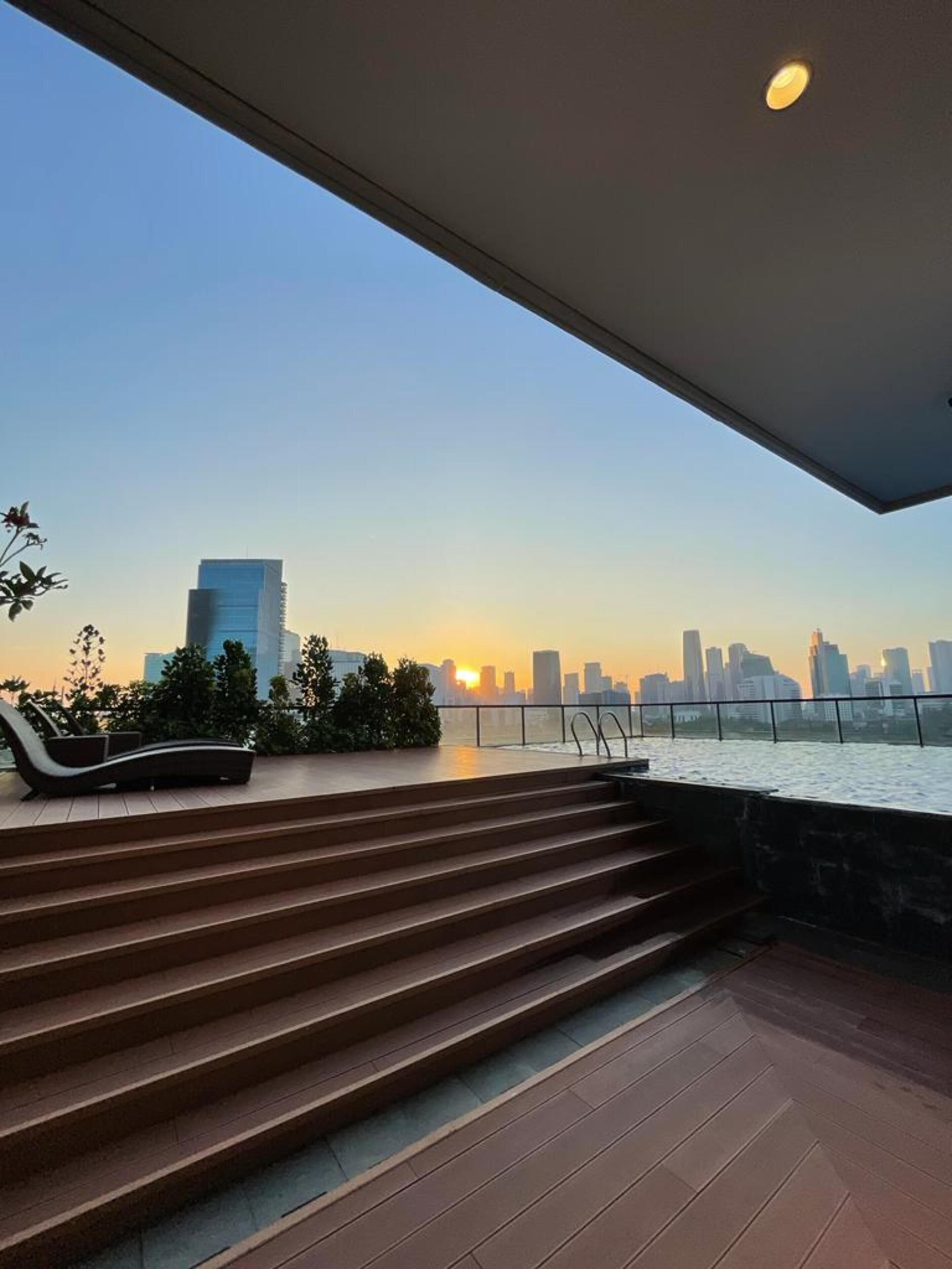 A modern outdoor area with a view of a city skyline at sunset, featuring a set of stairs leading to a seating area with a railing overlooking a body of water.