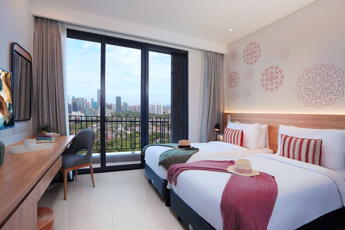 Ein Hotelzimmer mit großem Fenster, einem Schreibtisch, einem Bett mit Kissen und Blick auf die Stadt.