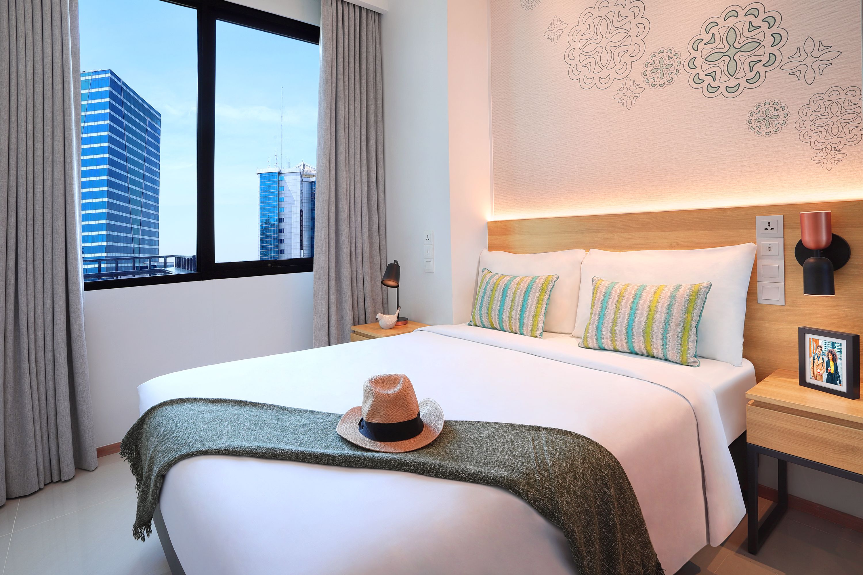 A hotel room with a large bed, a nightstand, a lamp, and a view of city buildings through the window.