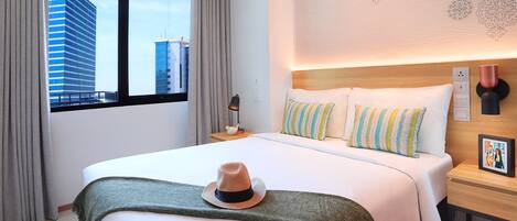A hotel room with a large bed, a nightstand, a lamp, and a view of city buildings through the window.