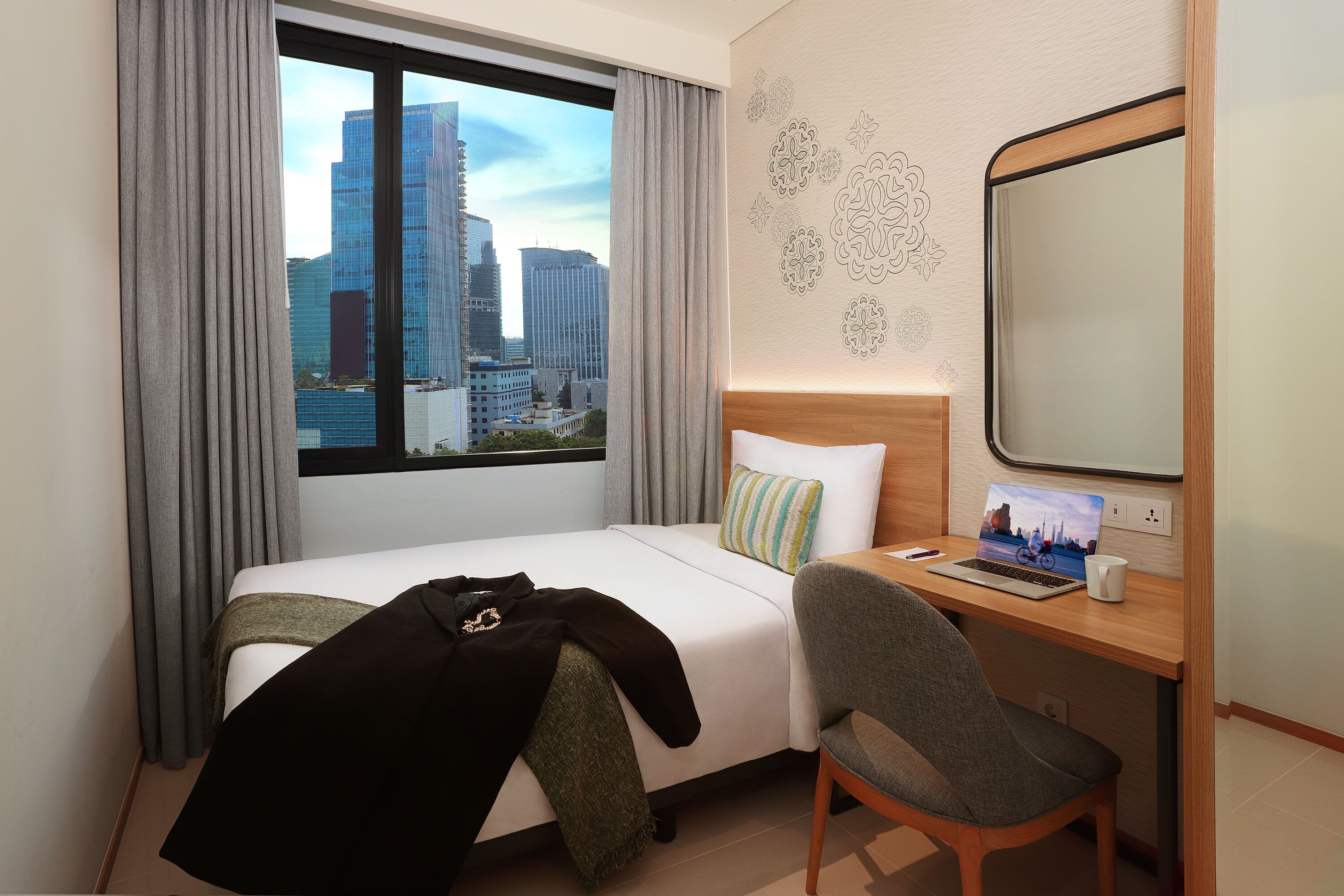 A hotel room with a bed, a desk with a laptop, a chair, and a large window offering a city view.
