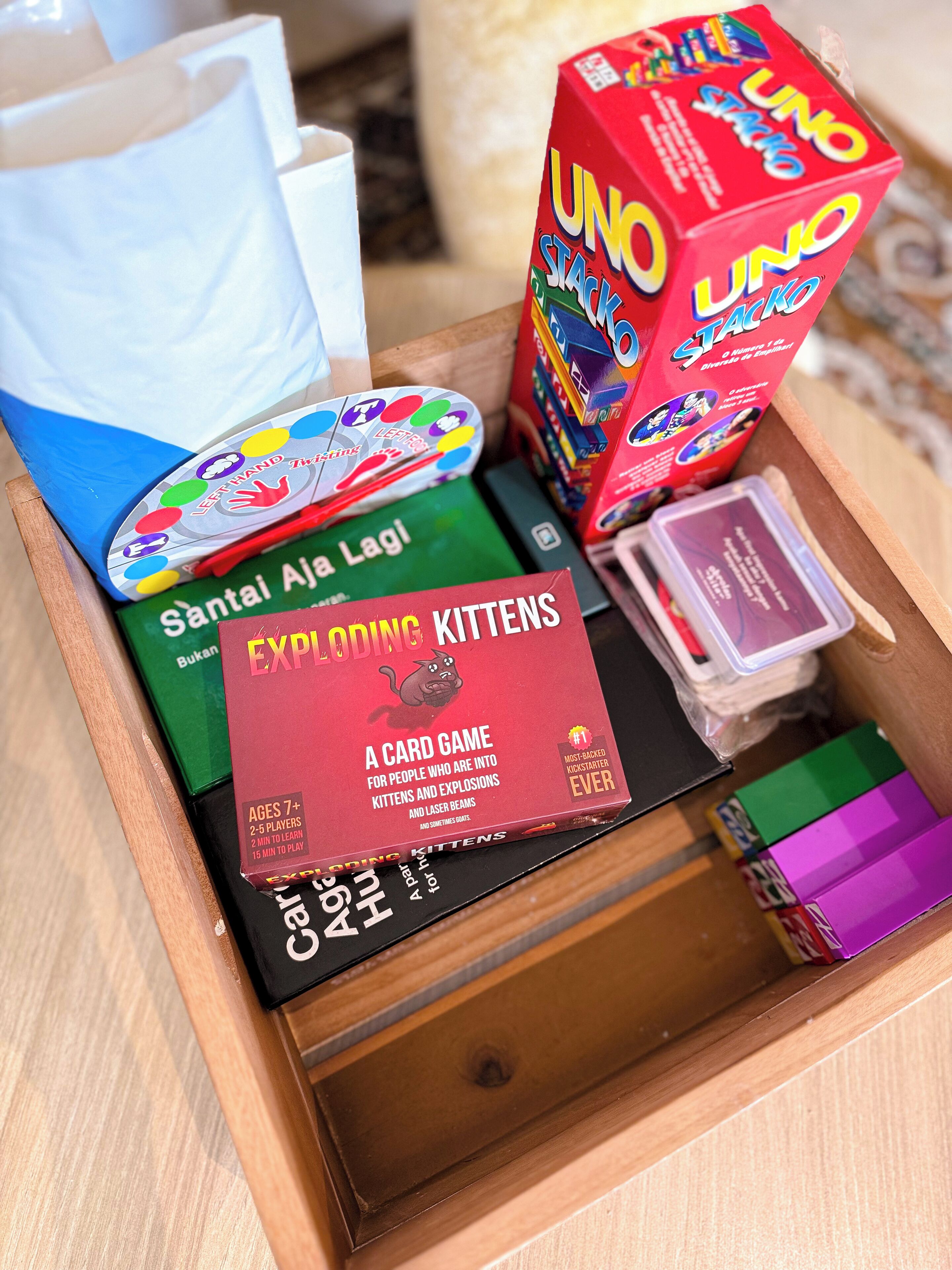 A wooden drawer containing various board games including UNO, Exploding Kittens, and a game called Santai A La Laga.