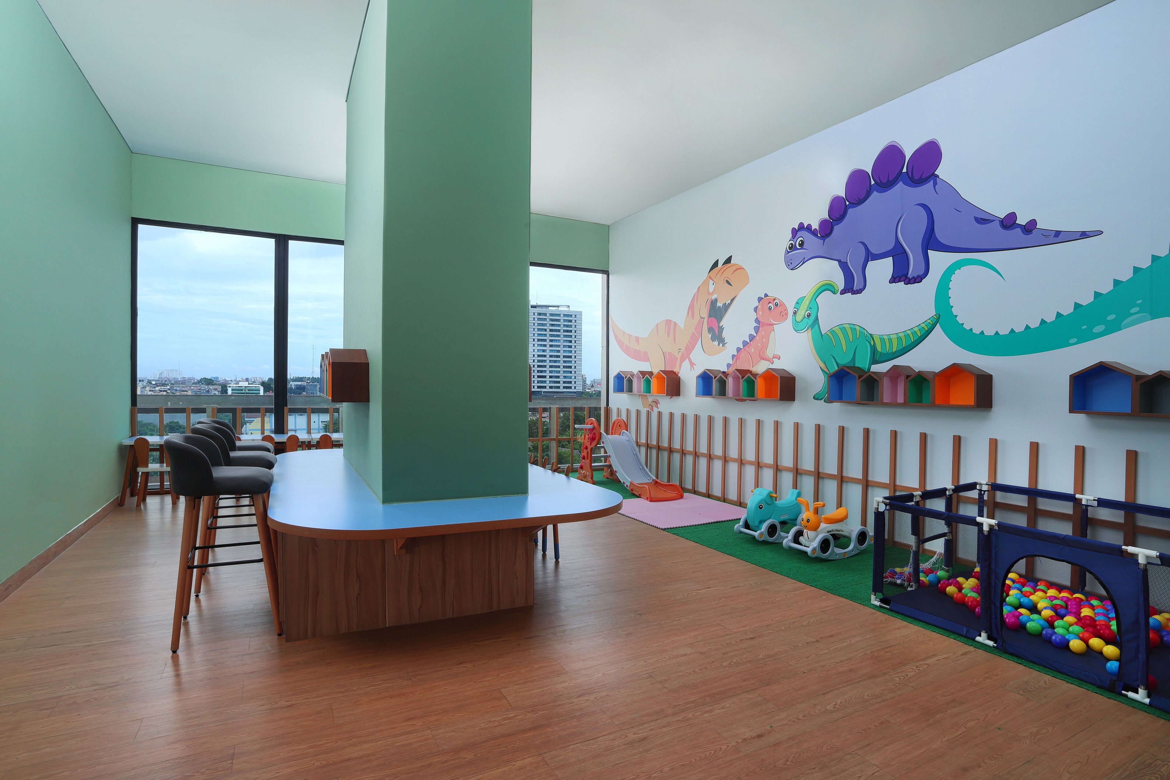 Zona de juegos para niños con un mural de dinosaurios, un corralito y vistas a la ciudad.