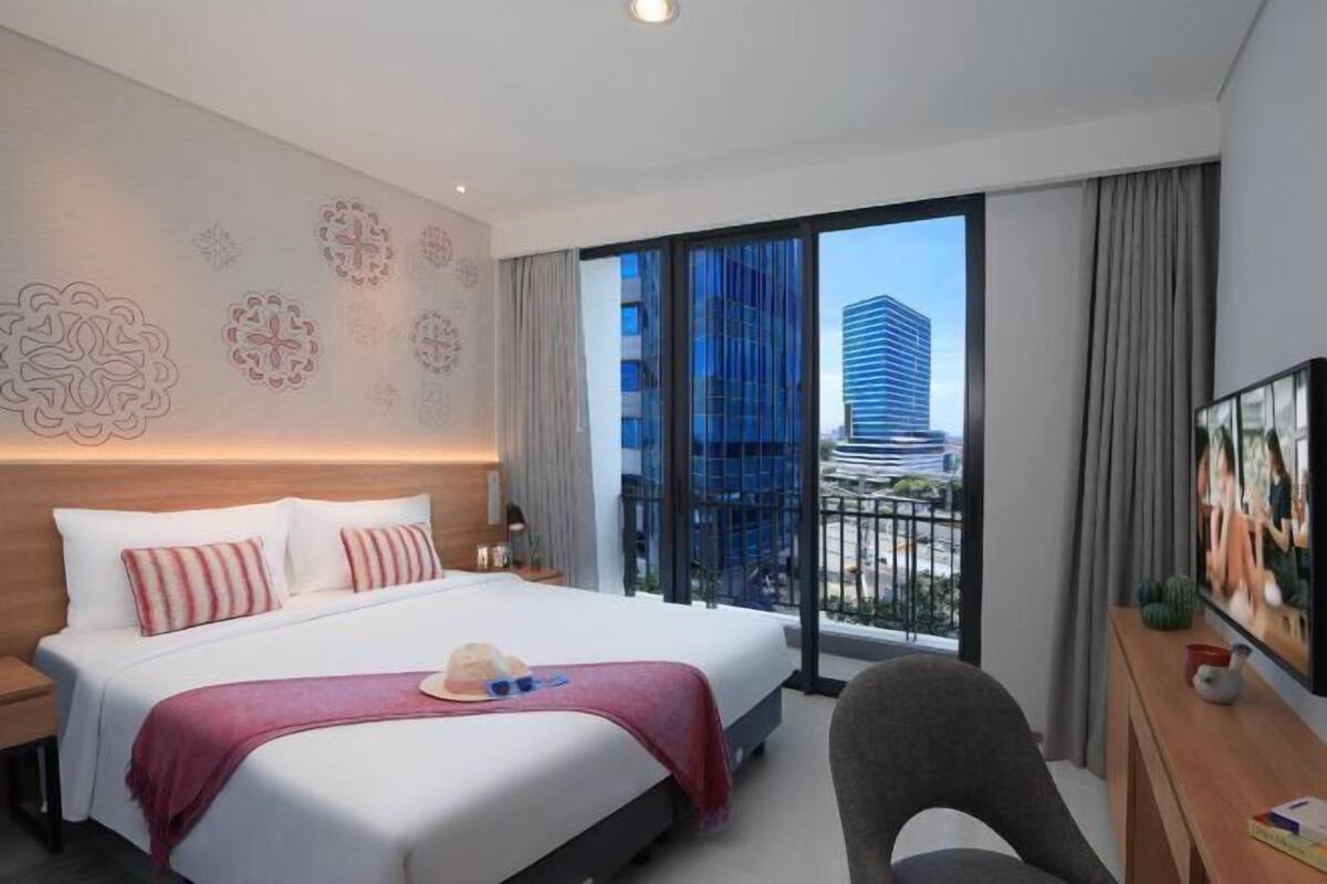 Ein Hotelzimmer mit einem großen Bett, einem Schreibtisch, einem Sessel, einem Fernseher und einem Balkon mit Blick auf die Stadt.