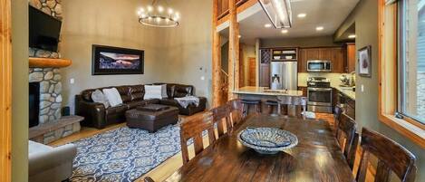 A spacious living room with a large wooden dining table, a fireplace, and a kitchen area visible through an open door.
