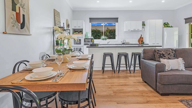 Una cocina moderna con armarios blancos, un comedor con mesa de madera y sillas negras, y un sofá gris con un cojín estampado.