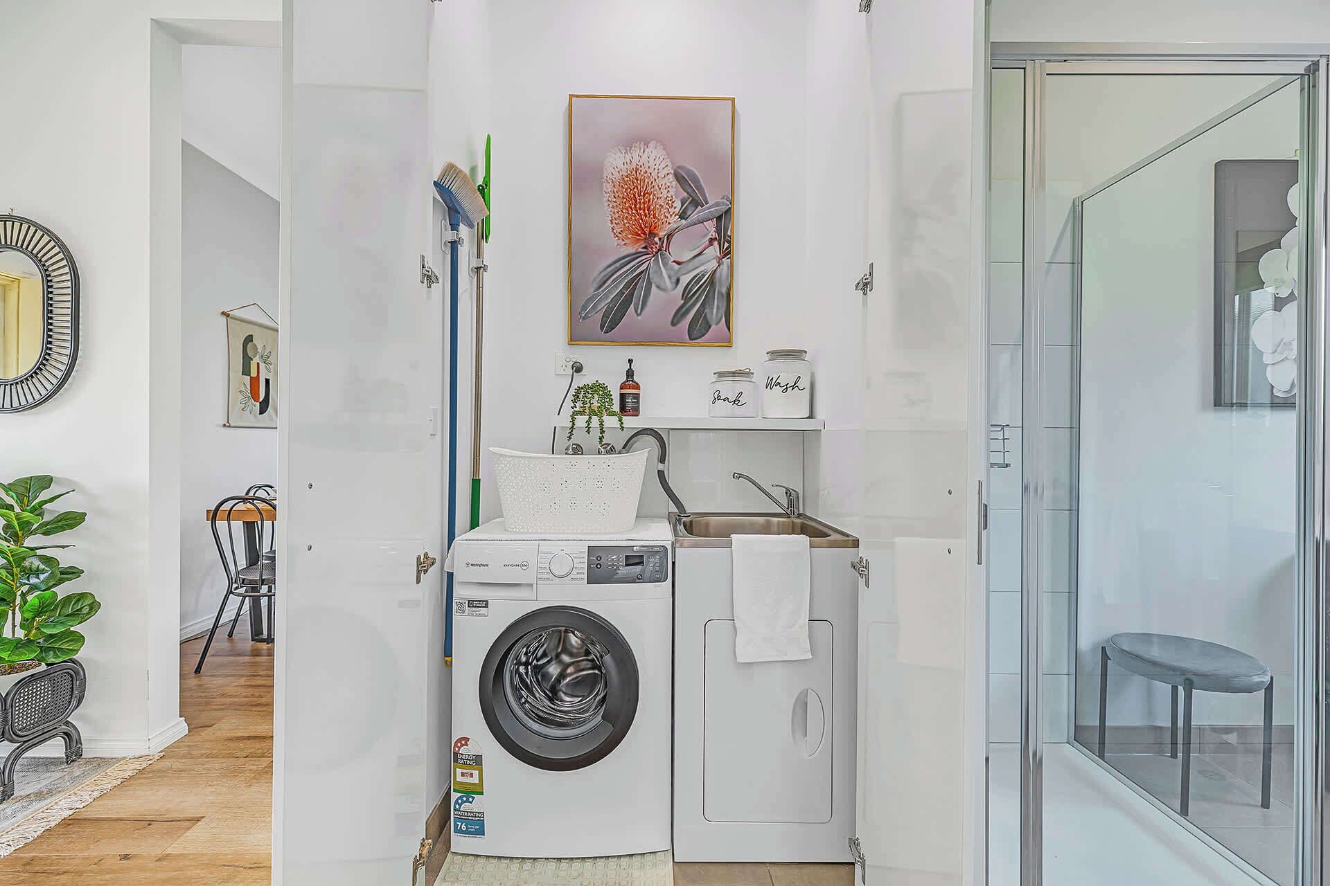 A compact laundry room with a front-loading washing machine, a sink, and a glass shower enclosure.