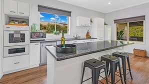 Una cocina moderna con muebles blancos, una isla grande con taburetes, horno integrado y vistas a un jardín a través del ventanal.