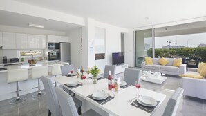 Cozinha moderna com armários brancos, área de jantar com mesa branca posta para quatro pessoas e sofá com almofadas amarelas.
