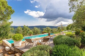 A pool area with a view of the countryside, surrounded by greenery and a cloudy sky.