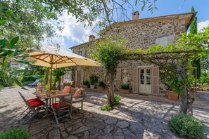 A stone building with a patio area featuring a table and chairs under a canopy, surrounded by potted plants and a pergola.