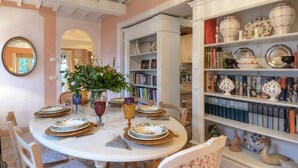 A dining area with a round table set for four, surrounded by chairs. There are bookshelves filled with books and decorative items. - Podere San Giorgio 6 Emma Villas (Manciano)
