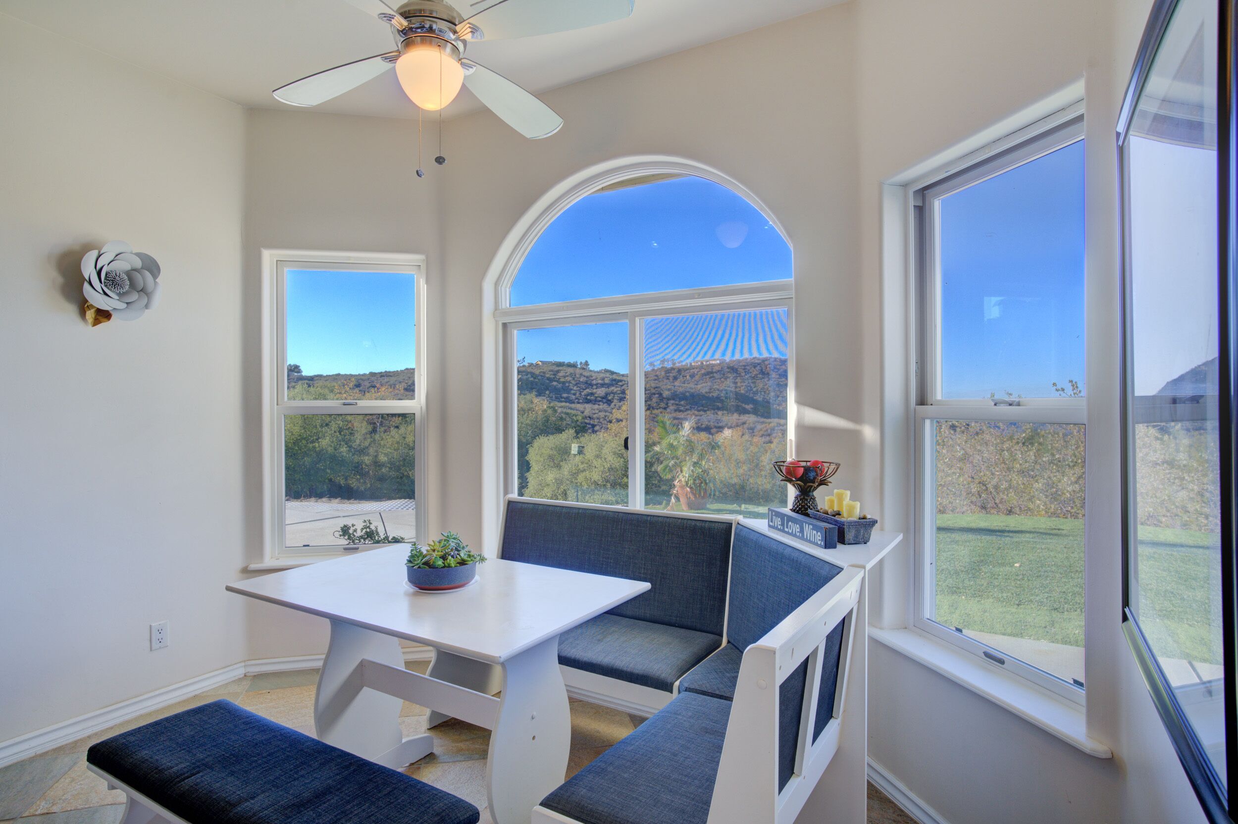 A room with a white table, blue bench seating, and large windows offering a view of a hilly landscape.