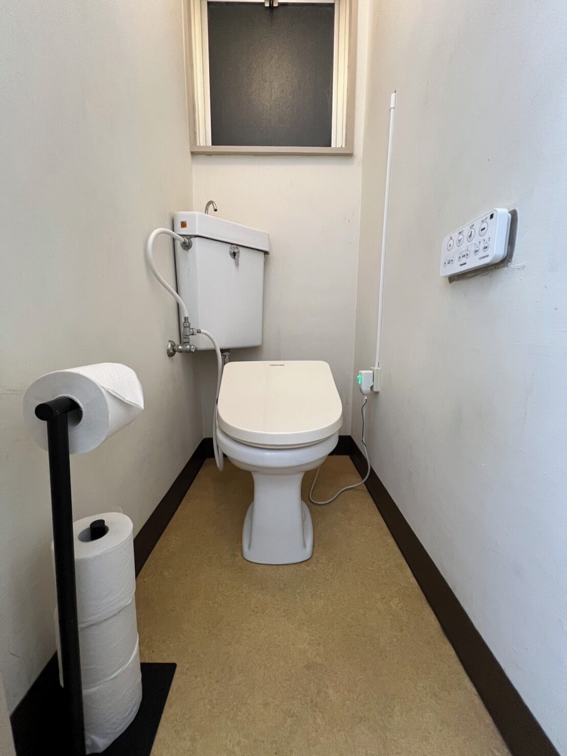 A small, clean bathroom with a toilet, a toilet paper holder, a waste bin, a window, and a wall-mounted electrical outlet.