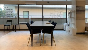 Un salón de conferencias con una mesa grande y sillas, ventanas amplias con vistas a edificios y árboles, y una escalera apoyada en la pared.
