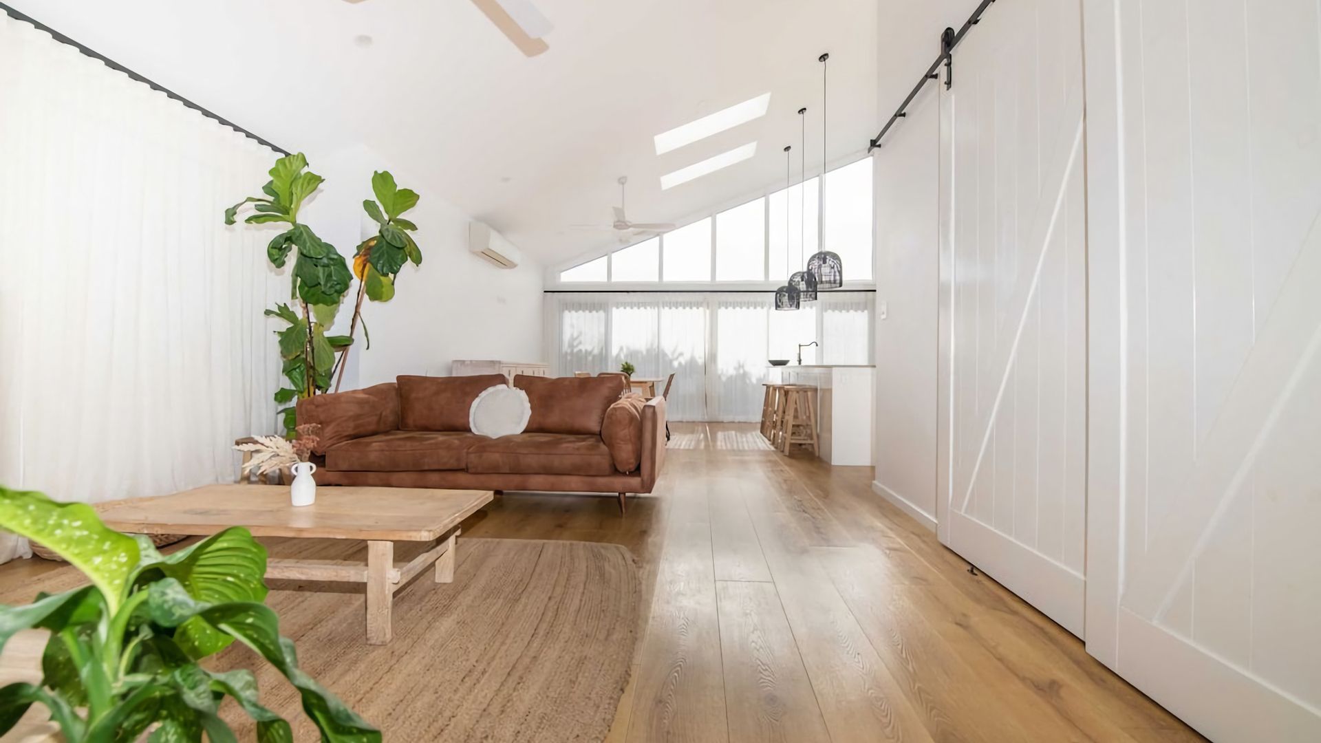 A spacious living area with a brown sofa, wooden flooring, and large windows allowing natural light.