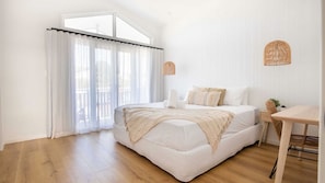 A bedroom with a bed, a wooden table, and a window with sheer curtains. - Surfleet Serenity is a coastal duplex (Kiama)