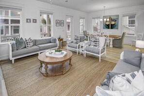 A spacious living room with a grey sofa, a round wooden coffee table, and a dining area with a wooden table and white chairs.