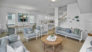 A spacious living room with a large gray sofa, a wooden coffee table, and a dining area with white chairs and a kitchen in the background. - Private *Heated Pool | Free Golf Cart | New Home | Patio Area | 5 Outdoor Spaces | Game Table (Santa Rosa Beach)