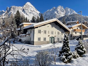 Un edificio cubierto de nieve con techo inclinado, una puerta verde y una vista despejada de montañas nevadas al fondo.