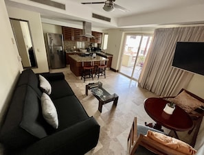 A modern living room with a black sofa, a coffee table, and a TV on a stand. There is a kitchen area with a stove, sink, and cabinets. A balcony with a sliding door is visible. - Condominio Luxury Puerto Vallarta, una Cuadra de la Playa y Malecón! (Puerto Vallarta)