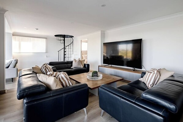 A modern living room with a black leather sofa set, a coffee table with decorative items, a flat-screen TV mounted on the wall, and a spiral staircase in the background. - Pia's Paradise Kiama (Kiama)
