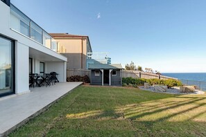 A modern house with a large glass window, a patio with outdoor furniture, and a view of the ocean.