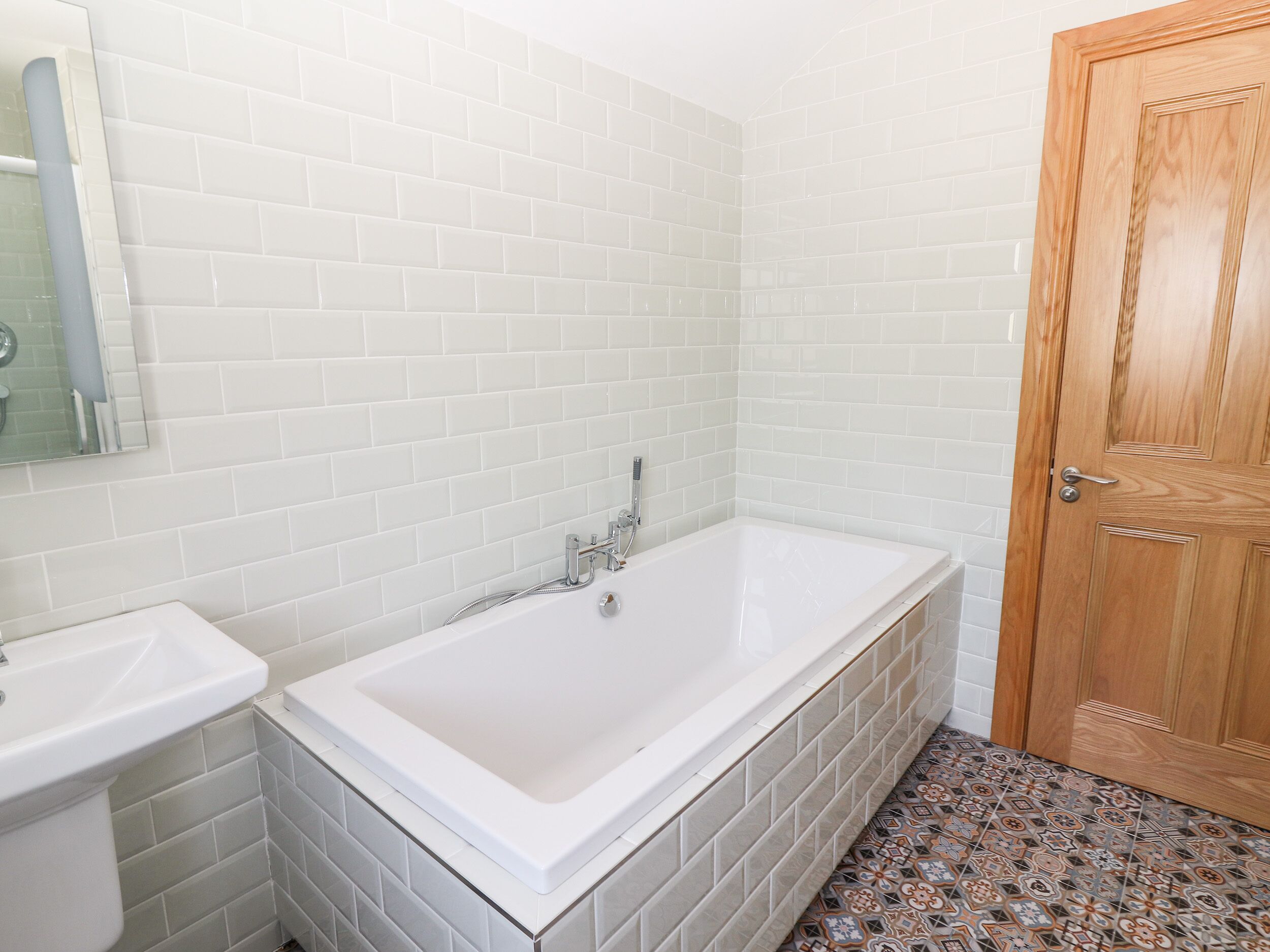 A bathroom with white tiled walls, a white bathtub, a sink, and a wooden door.