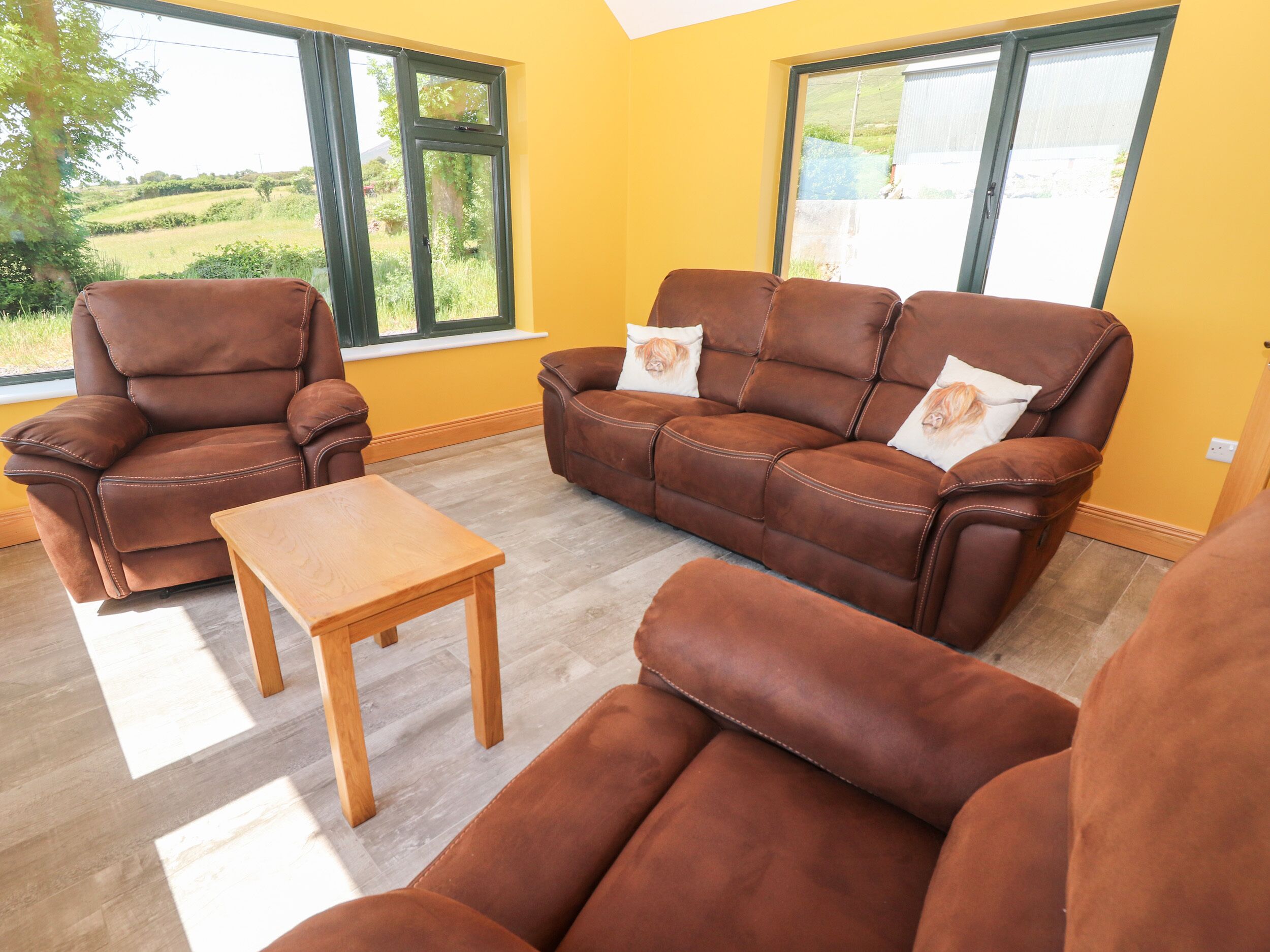 Ein Wohnzimmer mit braunen Ledersofas, einem Holztisch und großen Fenstern mit Blick auf Grünflächen.