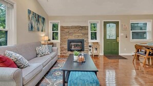 A spacious living room with a stone fireplace, a grey sofa, a coffee table, and a dining area with a wooden table and chairs.