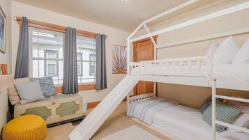 Een slaapkamer met een stapelbed en een houten ladder, een zithoek met kussens, en een raam met gordijnen.