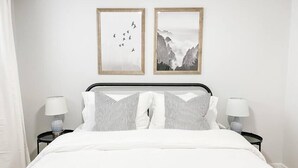 Un lit bien fait, avec une literie blanche et des oreillers à rayures, flanqué de deux tables de chevet surmontées de lampes, dans une pièce aux murs blancs et où deux tableaux encadrés sont accrochés au-dessus du lit.