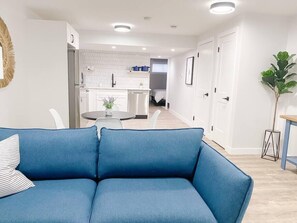 A modern living room with a blue sofa, a dining table with chairs, and a kitchen in the background. - Paisley Suite. Dog Friendly. (Edmonton)