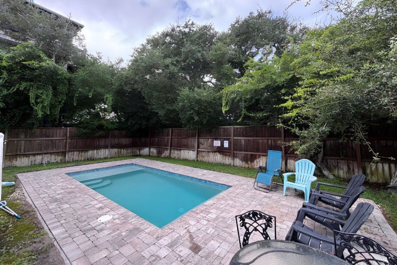 Une piscine rectangulaire, bordée d’une terrasse carrelée et entourée d’une clôture en bois et d’arbres.